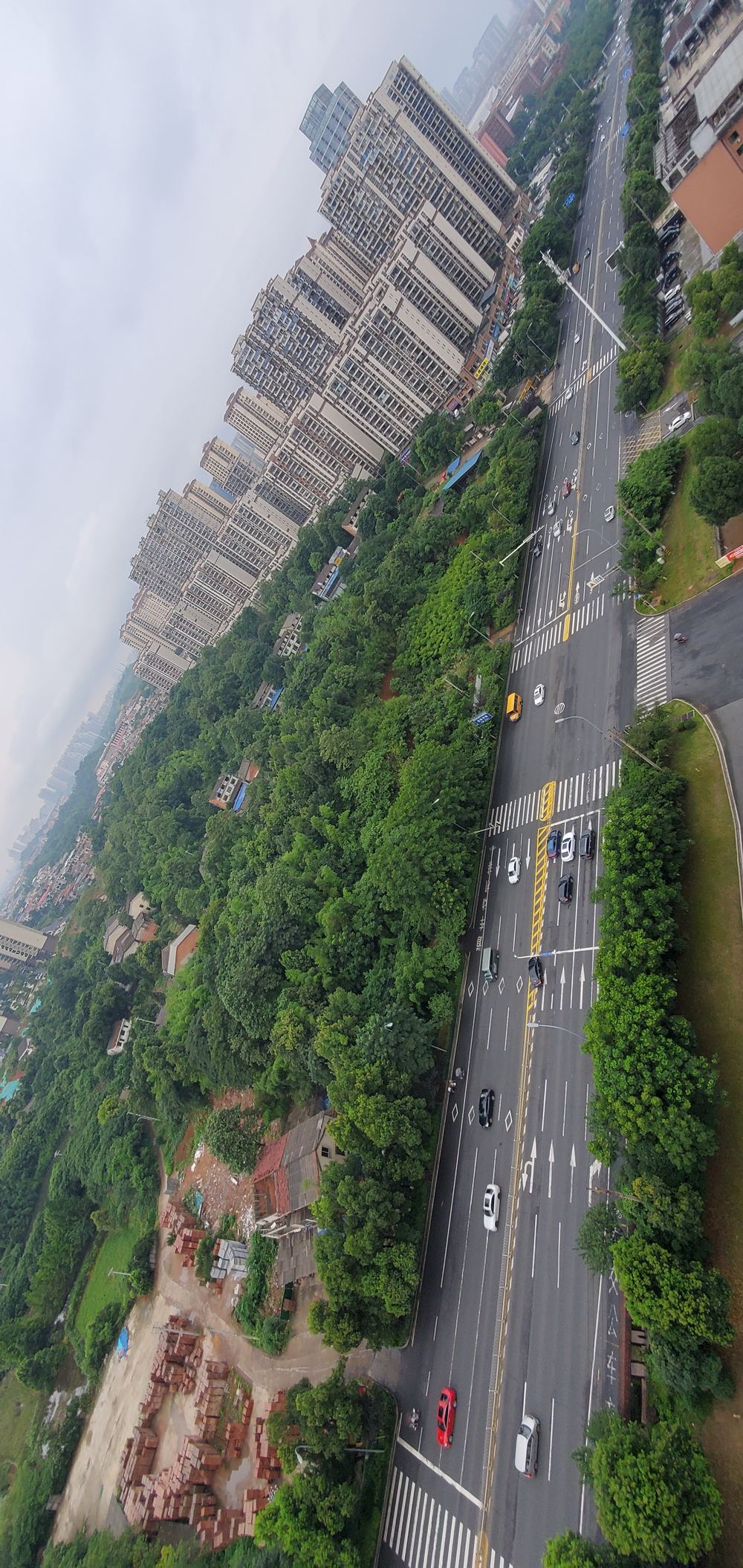 Aerial view from a 21st-floor apartment balcony in Yiyang, China, looking down at a tree-lined boulevard, moving traffic, and residential high-rises in the distance